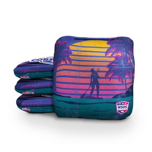 Beach cornhole bags beside a regulation board in a coastal sunset setting
